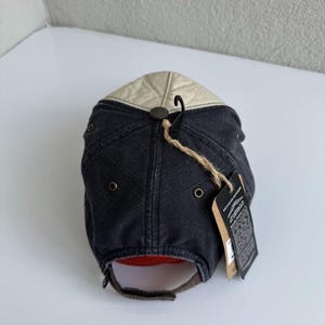 K&ouml;nnte beinhalten: Eine Baseballkappe mit dunkelanthrazitfarbener Krone, einem dezenten diagonalen Gittermuster und einem kontrastierenden cremefarbenen Oberteil. Die Kappe verf&uuml;gt &uuml;ber Metall&ouml;sen, einen braunen Stoffriemenverschluss und ein h&auml;ngendes Kenneth Howard-Markenetikett.