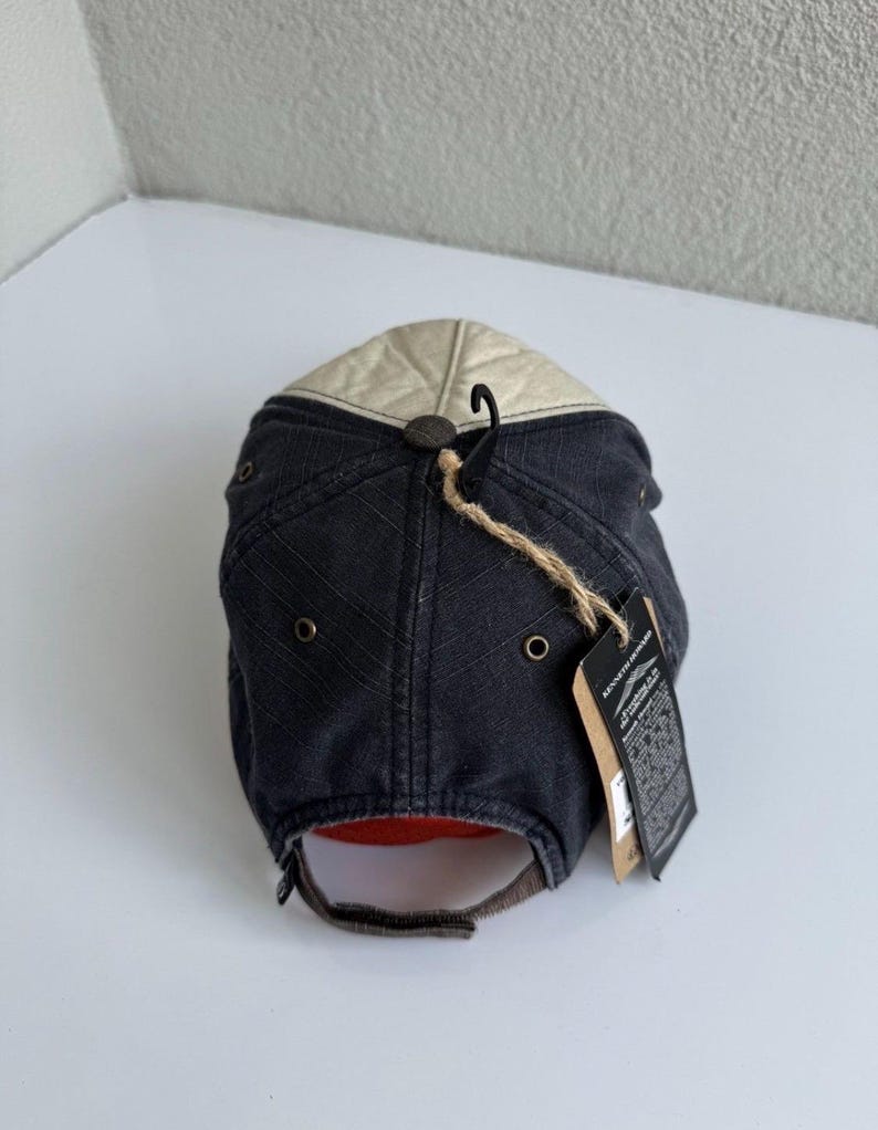 K&ouml;nnte beinhalten: Eine Baseballkappe mit dunkelanthrazitfarbener Krone, einem dezenten diagonalen Gittermuster und einem kontrastierenden cremefarbenen Oberteil. Die Kappe verf&uuml;gt &uuml;ber Metall&ouml;sen, einen braunen Stoffriemenverschluss und ein h&auml;ngendes Kenneth Howard-Markenetikett.
