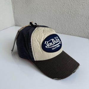 K&ouml;nnte beinhalten: Eine Baseballkappe mit dunkelblauer R&uuml;ckseite, cremefarbenem Vorderteil und dunkelbraunem Schirm mit abgenutzten Kanten. Ein dunkelblauer, ovaler Aufn&auml;her auf der Vorderseite zeigt den wei&szlig;en, gestickten Schriftzug Von Dutch California.