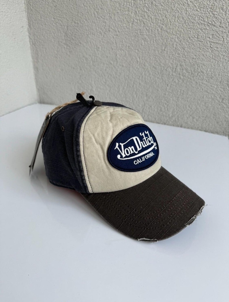K&ouml;nnte beinhalten: Eine Baseballkappe mit dunkelblauer R&uuml;ckseite, cremefarbenem Vorderteil und dunkelbraunem Schirm mit abgenutzten Kanten. Ein dunkelblauer, ovaler Aufn&auml;her auf der Vorderseite zeigt den wei&szlig;en, gestickten Schriftzug Von Dutch California.
