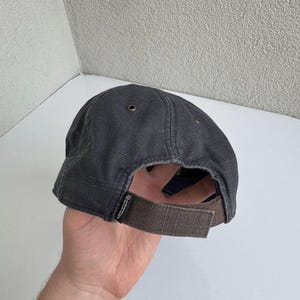 K&ouml;nnte beinhalten: Eine anthrazitfarbene Baseballkappe mit strukturierter Stoffoberfl&auml;che, einem braunen, verstellbaren Klettverschluss am Hinterkopf und kleinen Metall&ouml;sen an der Kappe. Ein kleines schwarzes Etikett mit wei&szlig;em Text ist an der Basis des Riemens befestigt.