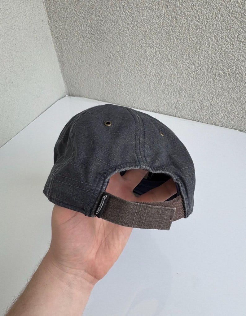 K&ouml;nnte beinhalten: Eine anthrazitfarbene Baseballkappe mit strukturierter Stoffoberfl&auml;che, einem braunen, verstellbaren Klettverschluss am Hinterkopf und kleinen Metall&ouml;sen an der Kappe. Ein kleines schwarzes Etikett mit wei&szlig;em Text ist an der Basis des Riemens befestigt.