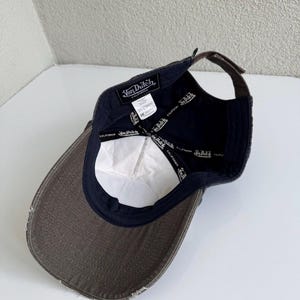 K&ouml;nnte beinhalten: Eine dunkelgraue Baseballkappe mit abgenutztem Schirm, von innen betrachtet. Das Innenfutter zeigt ein schwarzes Band mit dem wiederholt aufgedruckten Von Dutch California Logo. Ein wei&szlig;es Pflegeetikett und ein schwarzes Markenetikett sind sichtbar.