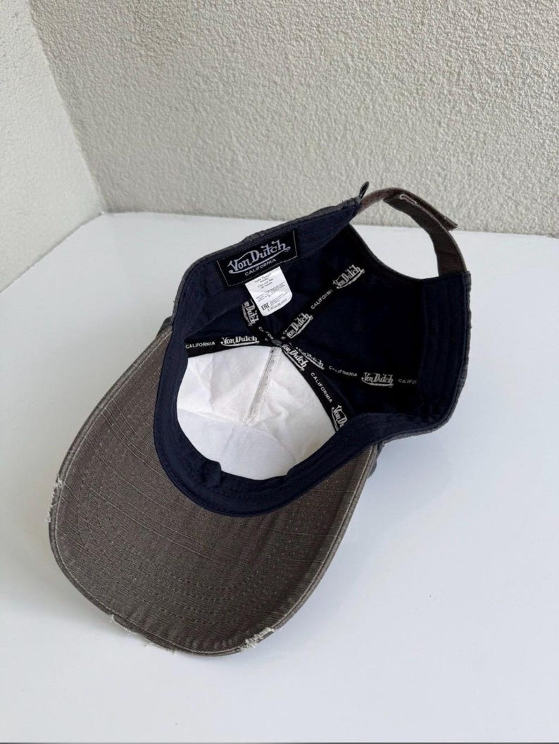 K&ouml;nnte beinhalten: Eine dunkelgraue Baseballkappe mit abgenutztem Schirm, von innen betrachtet. Das Innenfutter zeigt ein schwarzes Band mit dem wiederholt aufgedruckten Von Dutch California Logo. Ein wei&szlig;es Pflegeetikett und ein schwarzes Markenetikett sind sichtbar.