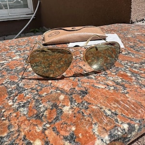 Op de afbeelding: Goudkleurige pilotenzonnebril met reflecterende, druppelvormige glazen en een dun metalen montuur. De zonnebril wordt vergezeld door een beige, lederachtig beschermend etui met een zichtbare logostempel op de voorflap.