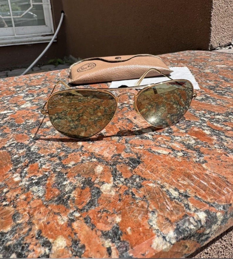 Op de afbeelding: Goudkleurige pilotenzonnebril met reflecterende, druppelvormige glazen en een dun metalen montuur. De zonnebril wordt vergezeld door een beige, lederachtig beschermend etui met een zichtbare logostempel op de voorflap.