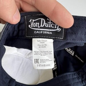 K&ouml;nnte beinhalten: Das Innere einer dunkelblauen Baseballkappe mit einem schwarzen rechteckigen Stoffetikett mit wei&szlig;em Von Dutch California Schriftzug. Ein wei&szlig;es Pflegeetikett zeigt die Texte Taille Unique Adulte, One size fits all Adults und 100% Coton.