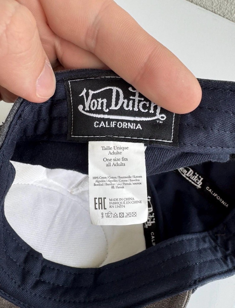 K&ouml;nnte beinhalten: Das Innere einer dunkelblauen Baseballkappe mit einem schwarzen rechteckigen Stoffetikett mit wei&szlig;em Von Dutch California Schriftzug. Ein wei&szlig;es Pflegeetikett zeigt die Texte Taille Unique Adulte, One size fits all Adults und 100% Coton.