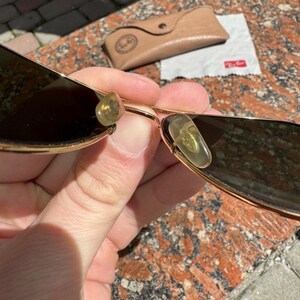 Op de afbeelding: Goudkleurige metalen zonnebril met donker getinte glazen en doorschijnende gele neuspads. De zonnebril wordt geleverd met een beige etui in lederlook en een klein wit reinigingsdoekje met het rode Ray-Ban-logo erop gedrukt.