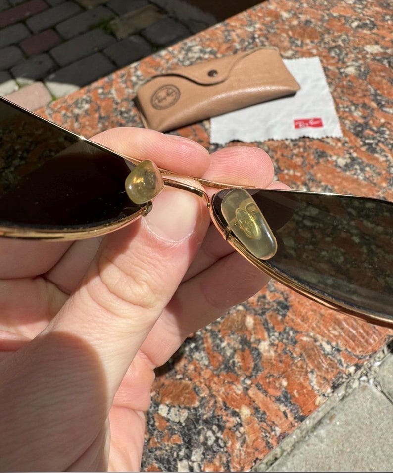 Op de afbeelding: Goudkleurige metalen zonnebril met donker getinte glazen en doorschijnende gele neuspads. De zonnebril wordt geleverd met een beige etui in lederlook en een klein wit reinigingsdoekje met het rode Ray-Ban-logo erop gedrukt.