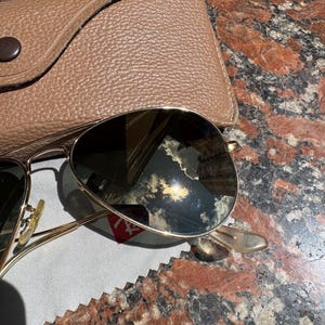 Op de afbeelding: Goudkleurige pilotenzonnebril met donkere, reflecterende glazen waarin de lucht en een gebouw te zien zijn. De zonnebril ligt naast een beige, gestructureerd lederen etui en een lichtgrijs reinigingsdoekje met een klein rood logo.