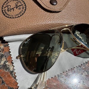 Op de afbeelding: Goudkleurige pilotenzonnebril met donkere glazen en het Ray-Ban-logo op het rechterbovenste glas. De zonnebril wordt geleverd met een beige, getextureerde lederen etui met drukknoopsluiting en een grijs microvezel reinigingsdoekje.