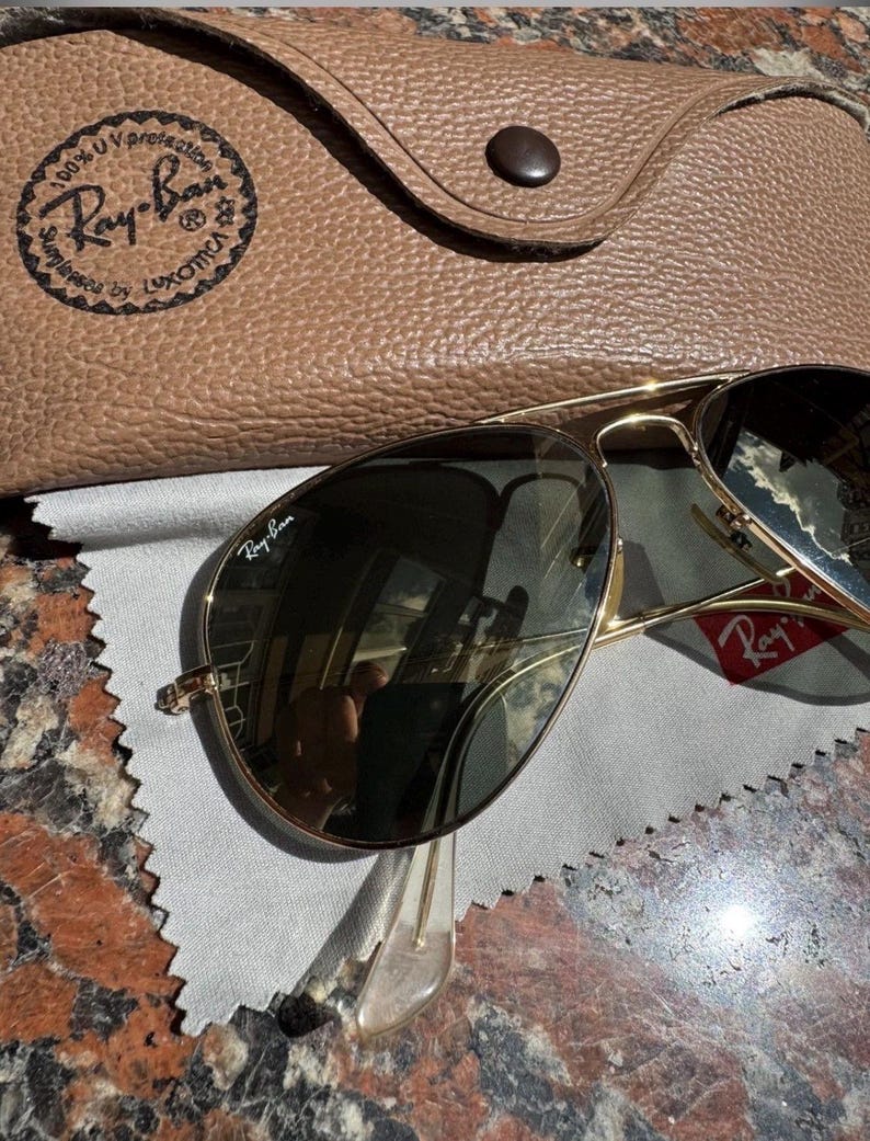 Op de afbeelding: Goudkleurige pilotenzonnebril met donkere glazen en het Ray-Ban-logo op het rechterbovenste glas. De zonnebril wordt geleverd met een beige, getextureerde lederen etui met drukknoopsluiting en een grijs microvezel reinigingsdoekje.