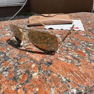 Op de afbeelding: Goudkleurige pilotenzonnebril met reflecterende druppelvormige glazen en een dun metalen montuur. Inbegrepen zijn een beige, gestructureerde etui in lederlook met drukknoopsluiting en een klein wit reinigingsdoekje met een rood logo.