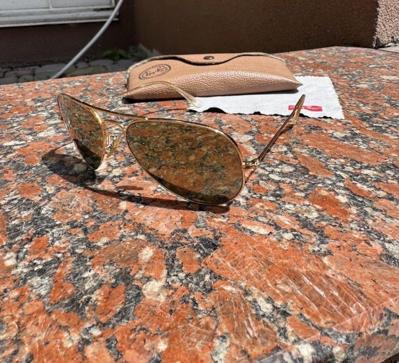 Op de afbeelding: Goudkleurige pilotenzonnebril met reflecterende druppelvormige glazen en een dun metalen montuur. Inbegrepen zijn een beige, gestructureerde etui in lederlook met drukknoopsluiting en een klein wit reinigingsdoekje met een rood logo.