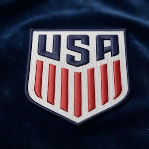 May include: An embroidered shield-shaped patch featuring the letters USA in dark blue above seven vertical red stripes on a cream-colored background. The patch is stitched onto a dark blue fabric surface with a soft, velvet-like texture.