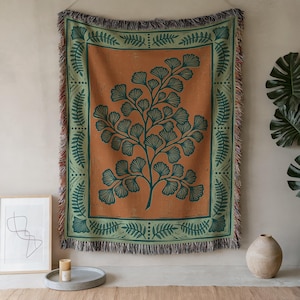 May include: A rectangular woven throw blanket featuring a central dark green ginkgo leaf branch design on a burnt orange background, framed by a decorative border of green fern leaves and geometric patterns with fringed edges on all sides.