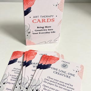 Peut inclure: Un jeu de cartes Art Therapy dans une boîte blanche avec des accents aquarelle rouges, noirs et roses. La boîte porte les inscriptions "OBSERVE CREATE EXPLOE", "ART THERAPY CARDS" et "Bring More Creativity Into Your Everyday Life".