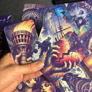 May include: A collection of rectangular tarot cards featuring vibrant, surreal illustrations including a flaming torch, a ship at sea, a cracked heart held by hands, and stylized portraits of people, all rendered in deep blues, purples, and warm oranges.
