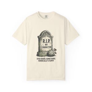 May include: A cream-colored short-sleeve t-shirt featuring a graphic of a tombstone with the text R.I.P. MY CHARACTER and a twenty-sided die at the base. Below the tombstone, the text reads DIED DOING SOMETHING HEROICALLY STUPID.