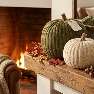 May include: Three ribbed knit pumpkins in olive green and cream colours, featuring a small tag on the green pumpkin with the text Cozy. The pumpkins are arranged with scattered autumn leaves, a stack of books, and a candle in a black holder.