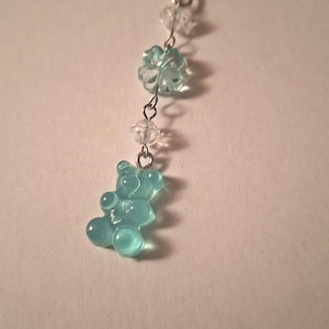 Könnte beinhalten: Ein Schlüsselanhänger mit einem silberfarbenen Metallring und einer Kette, verziert mit einem hellblauen, durchscheinenden Gummibärchen-Anhänger, einer kleinen facettierten transparenten Perle und einer hellblauen blütenförmigen Perle.