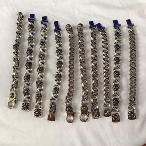 May include: A collection of ten silver-toned metal bracelets arranged in a row. Each bracelet features a chain-link design with various circular floral or cross-shaped charms, some incorporating blue accents and metallic clasps at the ends.