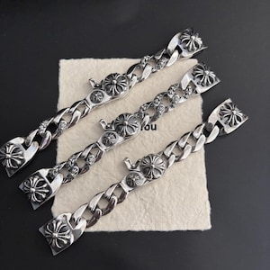 Può includere: Tre bracciali in metallo color argento con un design a catena a maglie spesse. Ogni bracciale include una chiusura circolare prominente decorata con un motivo a croce e ulteriori elementi decorativi a forma di croce integrati nelle maglie.