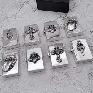 May include: A collection of seven rectangular silver-toned metal lighters arranged on a textured surface. Each lighter features a distinct raised emblem, including ornate crosses, fleur-de-lis motifs, and stylized lips with tongues.