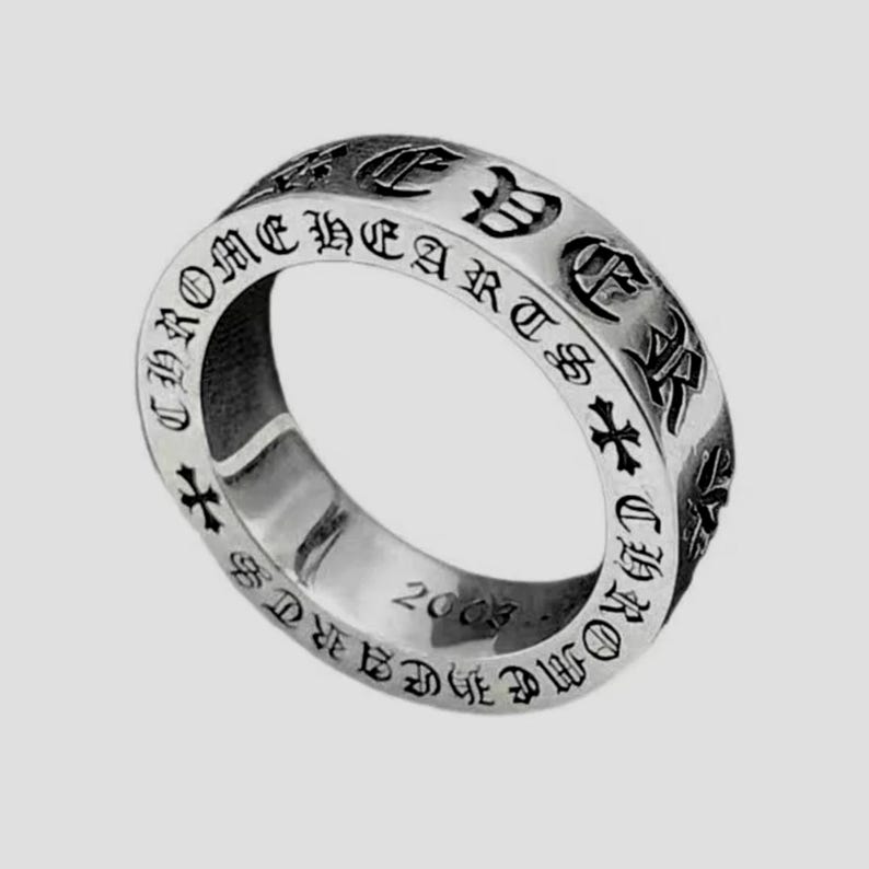 K&ouml;nnte beinhalten: Ein silberfarbener Metallring mit der eingravierten Aufschrift CHROME HEARTS in einer gotischen Schriftart auf dem &auml;u&szlig;eren Band. Das Design enth&auml;lt kleine Kreuzsymbole zwischen den Buchstaben und die Jahreszahl 2003 auf der Innenseite.