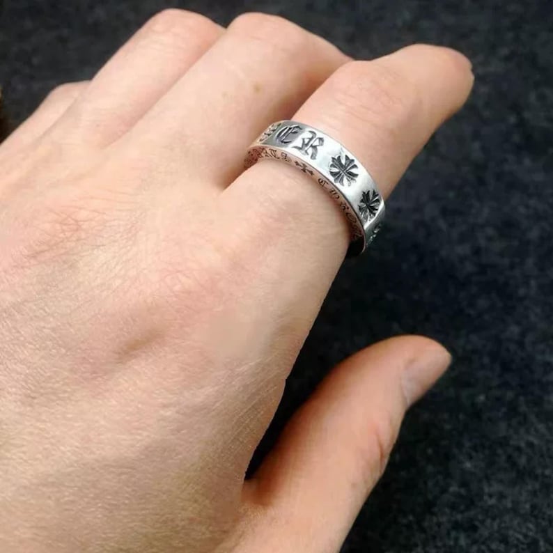 K&ouml;nnte beinhalten: Ein Ring aus silberfarbenem Metall mit einem Band, das mit gotischen Schriftzeichen und kleinen Kreuzsymbolen graviert ist. Das Band hat eine polierte Oberfl&auml;che mit abgedunkelten vertieften Bereichen, die die Gravuren hervorheben.