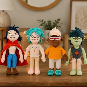May include: Four plush dolls standing in a row. The first has dark hair, a red cape, and blue pants. The second has teal hair and a white shirt. The third has a peach cap, orange shirt, and teal shoes. The fourth has green skin and a dark tank top.