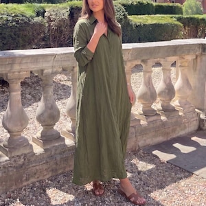 Könnte beinhalten: Ein langärmeliges olivgrünes Hemdkleid mit durchgehender Knopfleiste, klassischem Kragen und einer lockeren, fließenden Silhouette, die bis zu den Knöcheln reicht, kombiniert mit braunen Ledersandalen zum Hineinschlüpfen.