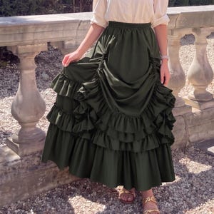 May include: A dark olive green maxi skirt featuring a high-waisted elastic band, vertical ruched detailing along the front, and multiple tiers of wide, gathered ruffles that create a voluminous, layered silhouette extending to the floor.