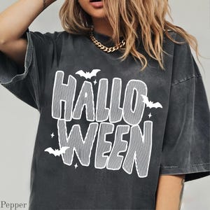 May include: A charcoal grey short-sleeve t-shirt featuring a white graphic print with the text "HALLOWEEN" in a striped font. The design includes small white bat silhouettes and star shapes surrounding the bold, vertically striped lettering.