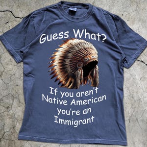 May include: A slate blue short-sleeve t-shirt featuring a graphic of a feathered headdress. The text above the graphic reads Guess What? and the text below reads If you aren't Native American You're an Immigrant in white lettering.