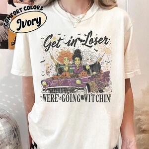 May include: An ivory short-sleeve t-shirt featuring a graphic of three witches in a purple convertible car with pumpkins. The text reads Get in Loser WE'RE GOING WITCHIN'. The design includes bats, stars, and fireworks, with a Comfort Colors Ivory label.