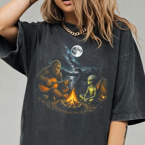 May include: A charcoal gray t-shirt featuring a graphic print of a Bigfoot playing an acoustic guitar by a campfire. Beside the fire sits a green alien, while a Loch Ness monster emerges from the water under a full moon and a starry night sky.