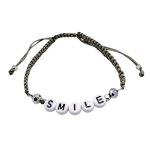 Op de afbeelding: Een armband met een gevlochten grijs koord en verstelbare trekkoorden. Het midden toont vijf witte ronde kralen, elk bedrukt met een zwarte letter om het woord SMILE te spellen. Twee metalen gefacetteerde kralen flankeren de kralen met letters.