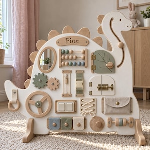 May include: A white dinosaur-shaped wooden busy board featuring the name Finn. It includes various interactive elements: an abacus, a mirror, gears, a zipper, a buckle, a latch, light switches, laces, a fabric pocket, and wooden rollers.