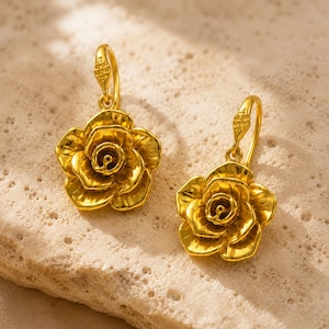 May include: A pair of gold-toned drop earrings featuring a three-dimensional rose flower design. Each earring has a curved hook wire with a small textured detail at the top, leading to a polished, layered petal rose bloom in a bright yellow gold finish.