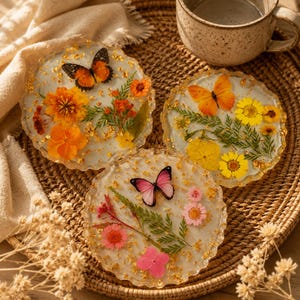 Puede incluir: Tres posavasos circulares de resina con flores secas, follaje verde y mariposas en naranja, amarillo y rosa. Cada posavasos tiene un borde festoneado y está decorado con copos de pan de oro incrustados en el material transparente.