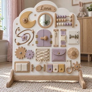 May include: A white wooden activity board featuring the name Luna at the top. It includes a moon shape, a circular mirror, an abacus with purple and beige beads, a small door, a zipper, a buckle, a metal latch, gears, laces, a pocket, and switches.