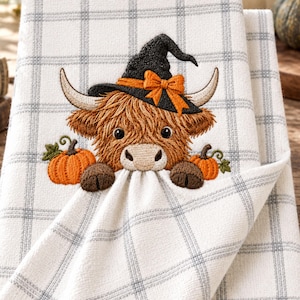 Puede incluir: Un paño de cocina a cuadros blancos y grises con un bordado de una vaca Highland marrón que lleva un sombrero de bruja negro con un lazo naranja. La vaca está situada entre dos pequeñas calabazas naranjas con tallos y hojas verdes.