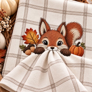 May include: A cream-colored kitchen towel featuring a brown plaid pattern and an embroidered design of a fox face with a bushy tail, a large orange maple leaf, two brown acorns, and a small orange pumpkin centered on the fabric surface.
