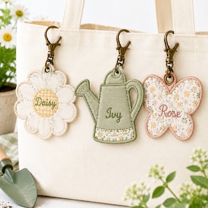 May include: Three fabric bag charms with bronze-toned lobster clasps attached to a cream canvas tote. The charms are shaped like a daisy with the name Daisy, a watering can with the name Ivy, and a butterfly with the name Rose embroidered on them.