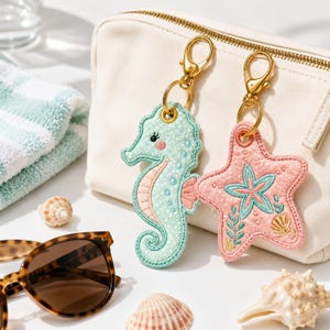 Puede incluir: Dos llaveros bordados con forma de caballito de mar verde menta y estrella de mar rosa, ambos unidos a mosquetones de metal dorado. El caballito de mar tiene lunares blancos y una mejilla rosa, mientras que la estrella de mar luce un motivo floral turquesa.