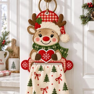 Puede incluir: Un soporte para toallas de cocina con un reno de peluche que lleva un gorro de cuadros rojos y blancos y una bufanda de cuadros verdes. El reno sostiene un corazón rojo con un copo de nieve blanco. Debajo cuelga una toalla crema con árboles.