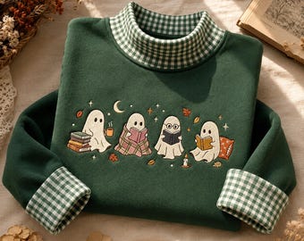 May include: A dark green long-sleeve sweatshirt featuring a mock neck collar and sleeve cuffs with a white and green gingham pattern. The chest displays an embroidered scene of four white ghosts reading books, drinking coffee, and sitting on pillows.