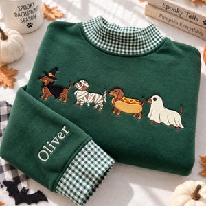 Puede incluir: Una sudadera de manga larga verde oscuro con cuello alto de patrón de cuadros vichy verde y blanco. El frente muestra teckels bordados disfrazados de bruja, momia, perrito caliente y fantasma. La manga está bordada con el nombre Oliver.